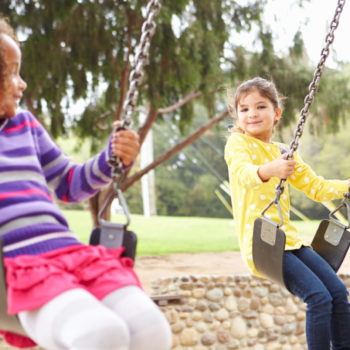 Two Young Girls Playing On Swing In Playground