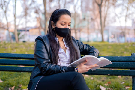 latina reading in park bench with face mask to prevent covid-19 coronavirus