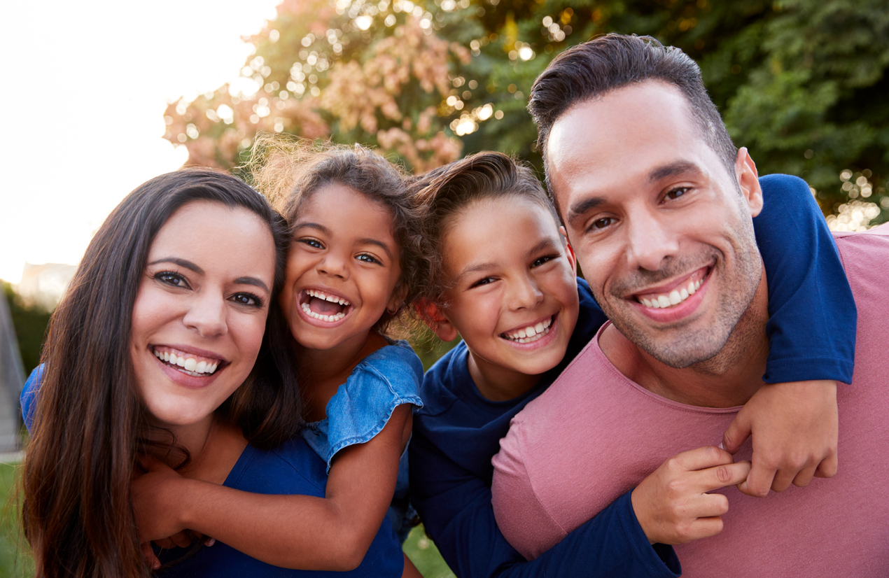 Portrait Of Smiling Hispanic Family With Parents Giving Children Piggyback Rides In Garden At Home