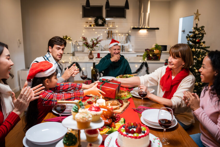 come home covered Multi-ethnic family exchanging presents during Christmas party at home. Young kid daughter receive gift from senior grandparent while having dinner to celebrate holiday Thanksgiving on dining table.