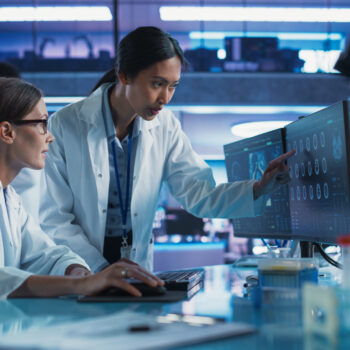 Female Doctors Talking In Medical Research Center And Using Desktop Computer To Analyze MRI Scans Of Brain. Surgeon And Neuroscientist Discussing Solutions For Treating Cancer.