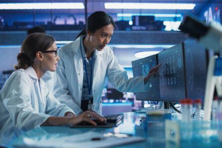 Female Doctors Talking In Medical Research Center And Using Desktop Computer To Analyze MRI Scans Of Brain. Surgeon And Neuroscientist Discussing Solutions For Treating Cancer.