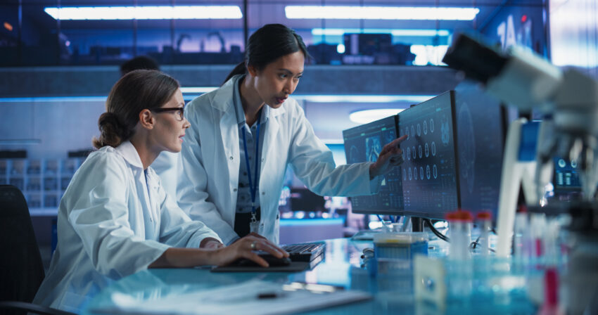Female Doctors Talking In Medical Research Center And Using Desktop Computer To Analyze MRI Scans Of Brain. Surgeon And Neuroscientist Discussing Solutions For Treating Cancer.