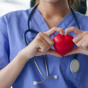 A nurse in blue scrubs holds a red heart in her hands, forming a heart shape around it, symbolizing care.