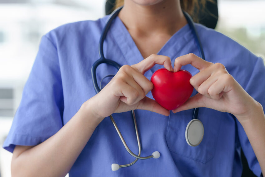 A nurse in blue scrubs holds a red heart in her hands, forming a heart shape around it, symbolizing care.