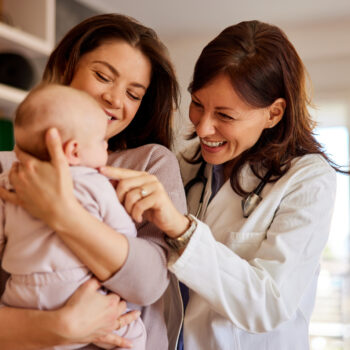 A smiling woman doctor pediatrician cuddles a cute baby girl on her cheek while her mother holding her.