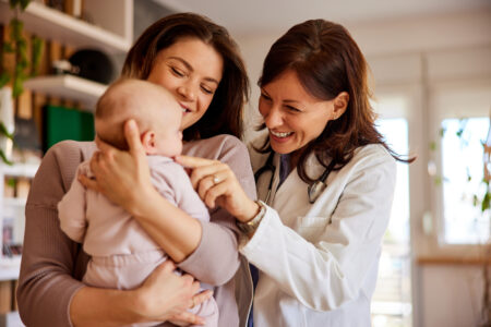 A smiling woman doctor pediatrician cuddles a cute baby girl on her cheek while her mother holding her.