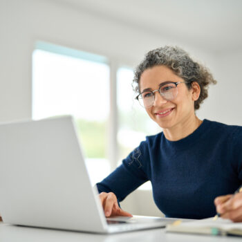 Happy middle aged woman sitting at table and using laptop writing notes.