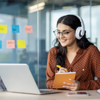 Woman watching online video course, training conference inside office at workplace. Office worker in headphones using laptop for remote meeting, writing data in notebook