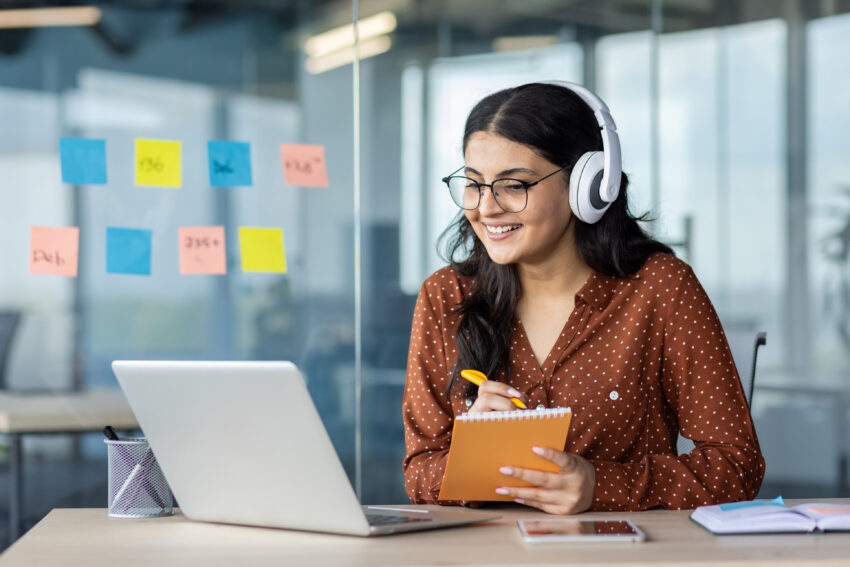 Woman watching online video course, training conference inside office at workplace. Office worker in headphones using laptop for remote meeting, writing data in notebook