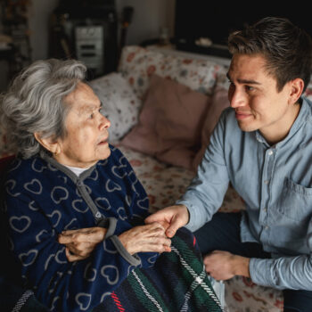 Young man sitting next to an old sick aged woman in wheelchair taking her hands while talking and smiling