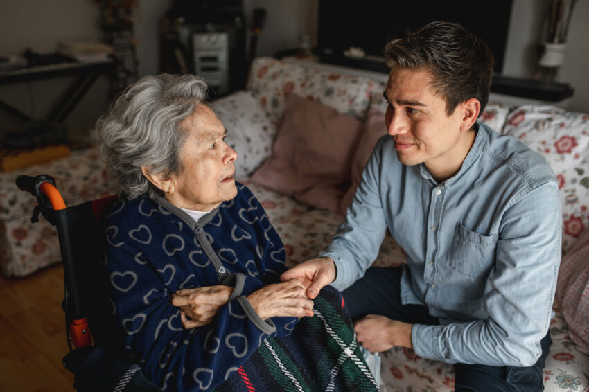 Young man sitting next to an old sick aged woman in wheelchair taking her hands while talking and smiling