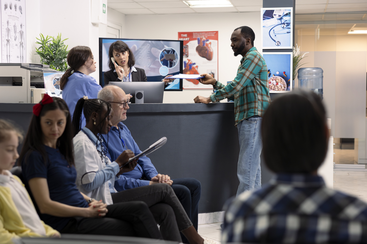 Hospital staff assists black man at reception desk while female doctor consults elderly male patient in clinic lobby. Receptionist multitasks with phone call and paperwork in busy waiting area.
