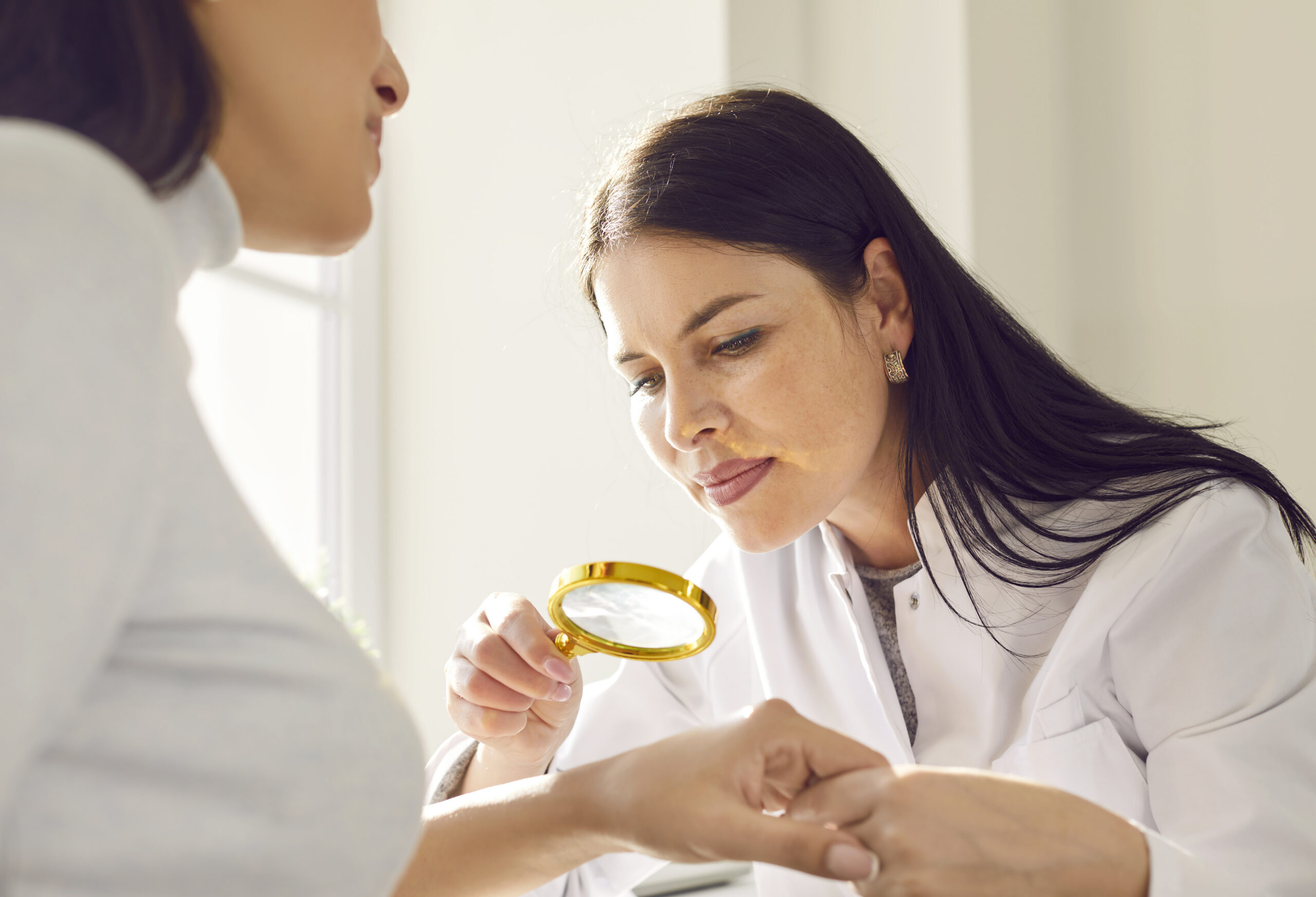 Female friendly doctor examining hand skin of young woman patient. Professional dermatologist using magnifying glass checking skin condition in clinic. Dermatology, skin health care concept.