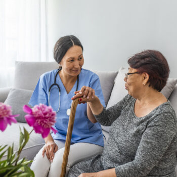 Close-up of elderly woman holding walking stick in the nursing house. Young carer supporting senior disabled woman with walking stick. Woman with walking stick assisted by young carer.