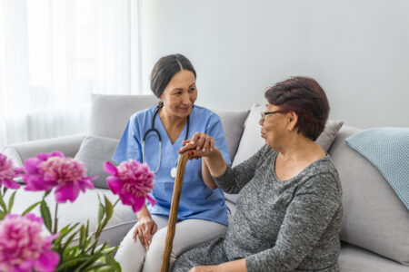 Close-up of elderly woman holding walking stick in the nursing house. Young carer supporting senior disabled woman with walking stick. Woman with walking stick assisted by young carer.