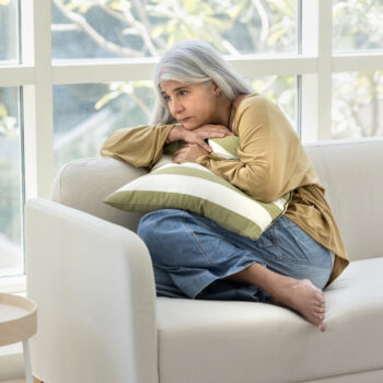 Sad depressed senior Hispanic lady hugging pillow on home couch, hugging pillow, keeping close pose, suffering from hard depression, memory disorder, anxiety, going through loss, grief. Full body shot