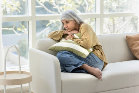 Sad depressed senior Hispanic lady hugging pillow on home couch, hugging pillow, keeping close pose, suffering from hard depression, memory disorder, anxiety, going through loss, grief. Full body shot